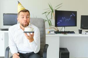 Jeune content homme d'affaire célébrer le sien anniversaire dans le bureau. il détient une gâteau dans le sien mains. photo