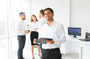 portrait de souriant Indien homme d'affaire permanent dans moderne Bureau sur collègues Contexte photo