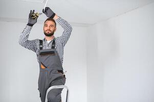 dépanneur ou professionnel électricien dans vêtements de travail installation lumière taches, permanent sur le échelle dans le blanc vivant pièce photo