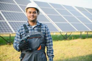portrait de Jeune Indien homme technicien portant blanc difficile chapeau permanent près solaire panneaux contre bleu ciel. industriel ouvrier solaire système installation photo
