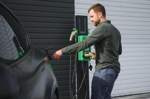 Beau barbu homme dans décontractée porter, permanent à le mise en charge station et chargeur pour un électrique auto. éco électrique voiture concept photo