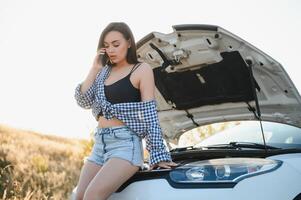 attrayant svelte Jeune fille dans été short et chemise réparations une cassé auto. une magnifique femme des stands près élevé voiture capot. photo