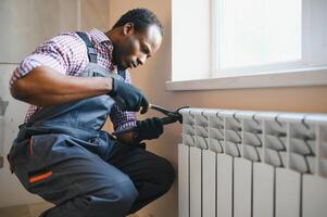 homme dans vêtements de travail salopette en utilisant outils tandis que installation ou réparer chauffage radiateur dans pièce photo