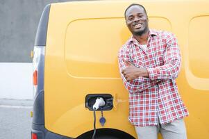 une africain américain homme des stands suivant à Jaune électrique livraison van à électrique véhicule mise en charge stations photo