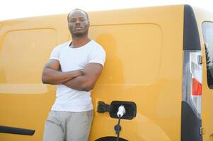 une africain américain homme des stands suivant à Jaune électrique livraison van à électrique véhicule mise en charge stations photo