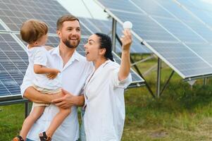 solaire énergie concept. une jeune, content famille est permanent près solaire panneaux et en portant un électrique lumière ampoule. photo