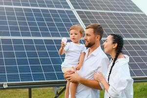 Jeune famille de Trois est accroupi près photovoltaïque solaire panneau, peu garçon et parents. moderne famille concept. le concept de vert énergie photo