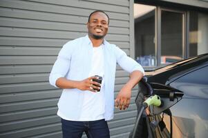 un Afro-américain homme est permanent près un électrique voiture, attendre pour il à charge à une mise en charge gare, et en buvant café. photo