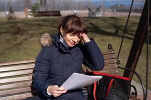 une Jeune femme comptable est assis sur une parc banc et lit un comptabilité document, agrippant sa diriger, rapports période photo