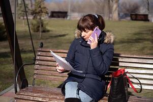 une Jeune femme comptable est assis sur une parc banc et lit une rapports document et appels sur une téléphone intelligent, rapports période photo