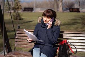 une Jeune femme comptable est assis sur une parc banc et lit une rapports document et appels sur une téléphone intelligent, rapports période photo