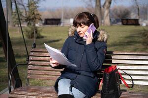 une Jeune femme comptable est assis sur une parc banc et lit une rapports document et appels sur une téléphone intelligent, rapports période photo