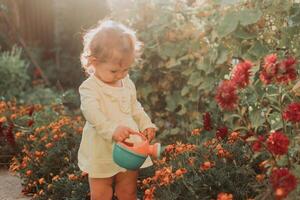 peu fille dans une Jaune robe et caoutchouc bottes est arrosage fleurs dans le l'automne jardin photo