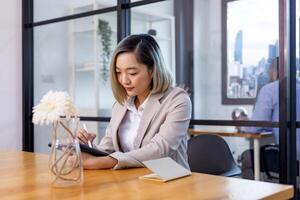 portrait de asiatique affaires PDG femme est travail dans Bureau à le table avec numérique tablette et montrant statistique graphique montrant annuel rapport et gratte-ciel Contexte photo