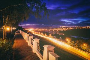 en mouvement voiture avec brouiller lumière par ville à nuit. pont plus de le rivière et le route. une vue de le parc de une la taille avec une clôture dans le premier plan. ancien film esthétique. photo