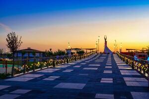 Ouzbékistan, Tachkent - avril 25, 2023 le territoire de le parc Nouveau Ouzbékistan avec monument de indépendance dans le forme de une stèle avec une humo oiseau à crépuscule. vue de une pont. photo