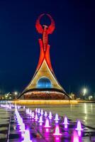 Ouzbékistan, Tachkent - mai 5, 2023 illuminé monument de indépendance dans le forme de une stèle avec une humo oiseau, fontaines et agitant drapeaux dans le Nouveau Ouzbékistan parc à la nuit. photo