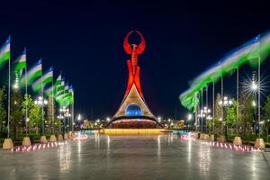 Ouzbékistan, Tachkent - mai 5, 2023 illuminé monument de indépendance dans le forme de une stèle avec une humo oiseau, fontaines et agitant drapeaux dans le Nouveau Ouzbékistan parc à la nuit. photo