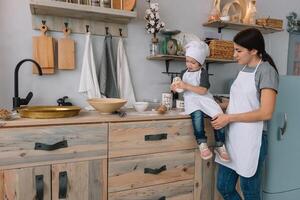 Jeune content maman et sa bébé cuisinier biscuits à Accueil dans le cuisine. Noël fait maison pain d'épice. mignonne garçon avec mère dans blanc uniforme et chapeau cuit Chocolat biscuits photo