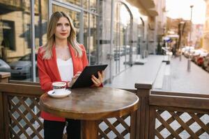 Extérieur ville mode portrait de Jeune femme d'affaires travail à café sur terrasse à ensoleillé jour, décontractée élégant tenue, menthe détails, en utilisant sa tablette, café casser, affaires concept photo