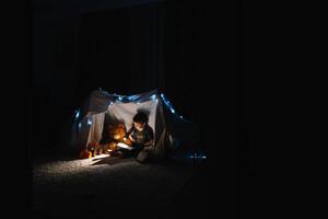 enfant garçon en train de lire avec livre et lampe de poche et nounours ours dans tente. avant Aller à lit. photo