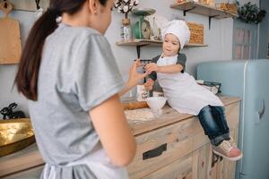 Jeune content maman et sa bébé cuisinier biscuits à Accueil dans le cuisine. Noël fait maison pain d'épice. mignonne garçon avec mère dans blanc uniforme et chapeau cuit Chocolat biscuits. photo