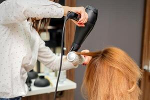 séchage cheveux dans le cheveux studio. femelle coiffeur styliste sèche cheveux avec une sèche-cheveux et rond brosse rouge cheveux de une femme dans une beauté salon. photo