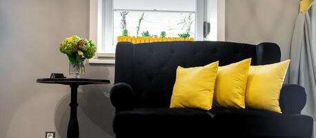 intérieur de le beauté chambre. noir canapé avec petit Jaune oreillers et café table sur une gris mur Contexte avec une la fenêtre. photo