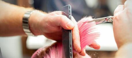 femme ayant une Nouveau la Coupe de cheveux. Masculin coiffeur Coupe rose cheveux avec les ciseaux dans une cheveux salon, proche en haut. photo