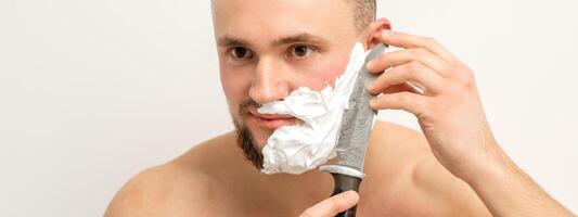 Jeune caucasien homme rasage barbe avec une gros couteau sur blanc Contexte. photo