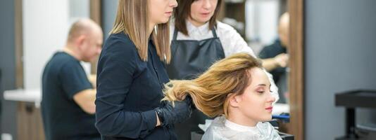 deux femelle les coiffeurs préparer longue cheveux de une Jeune femme fabrication boucles coiffure dans une beauté salon. photo