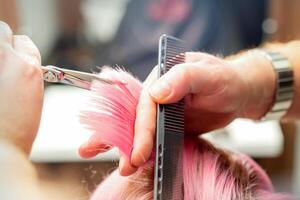 femme ayant une Nouveau la Coupe de cheveux. Masculin coiffeur Coupe rose cheveux avec les ciseaux dans une cheveux salon, proche en haut. photo