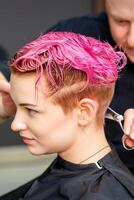 femme ayant une Nouveau la Coupe de cheveux. une Masculin coiffeur est Coupe teint rose court cheveux avec les ciseaux dans une cheveux salon. photo