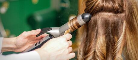 création boucles avec curling fers. coiffeur fait du une coiffure pour une Jeune femme avec longue rouge cheveux dans une beauté salon. photo