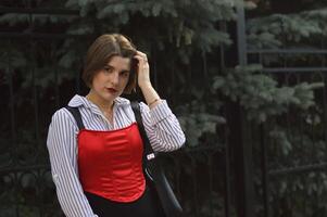 élégant attrayant femme dans une blanc chemise et rouge Haut contre une Contexte de à feuilles persistantes des arbres. ville portrait. photo