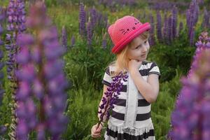 une blond fille dans une champ avec violet fleurs. une peu fille dans une rose chapeau est cueillette fleurs dans une champ. une champ avec lupins photo