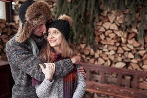 concept de noël et de couple - homme et femme souriants en chapeaux et écharpe étreignant sur une maison de campagne en bois et fond de neige photo