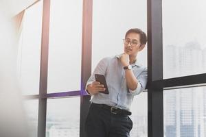 homme d'affaires asiatique debout à côté de la fenêtre en regardant le téléphone portable. photo