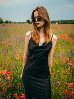 magnifique Jeune fille dans une noir soir robe et des lunettes de soleil posant contre une coquelicot champ sur une nuageux été journée. portrait de une femelle modèle en plein air. pluvieux temps. gris des nuages. photo