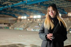 magnifique adolescent fille dans une noir hiver veste et chapeau pose contre le Contexte de une détruit et abandonné des sports salle. Urbain concept. photo