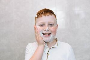 marrant garçon dans le une baignoire frottis le sien visage avec rasage crème, le enfant imite un adulte papa se rase. le garçon enduit le sien des joues avec rasage mousse photo