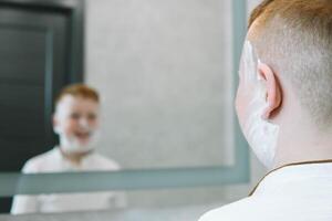 marrant garçon dans le une baignoire frottis le sien visage avec rasage crème, le enfant imite un adulte papa se rase. le garçon enduit le sien des joues avec rasage mousse photo