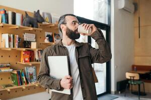 élégant et moderne barbu homme dans des lunettes permanent en buvant café dans une café magasin sur une ensoleillé jour, en portant une portable. pigiste photo