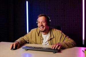Beau homme dans chemise vêtements et casque séance à une table dans une pièce avec néon lumières et en jouant Jeux sur le ordinateur avec une sourire sur le sien visage à la recherche à le filtrer. photo
