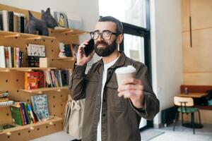 élégant barbu homme dans une chemise dans des lunettes des stands sur une Contexte de livres, en portant une tasse de café dans une café et souriant à le caméra. photo