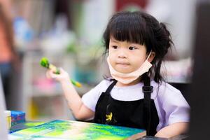 asiatique peu fille est séance en portant une grand pinceau et La peinture aquarelle sur une toile, enfant jouit apprentissage art, mignonne enfant loisir activité. les enfants vieilli 4 années. photo