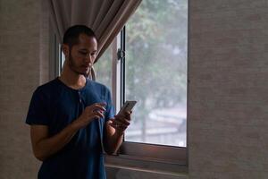 Jeune asiatique homme permanent sur fenêtre tandis que vérification le sien téléphone intelligent photo