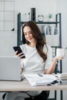 femme d'affaires chèques sa téléphone intelligent tandis que en portant une tasse de café, une portable ouvert sur le bureau dans de face de son. photo