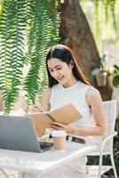 Jeune femme prend Remarques dans une spirale carnet à côté de sa portable à un Extérieur tableau, profiter une ensoleillé travail paramètre. photo