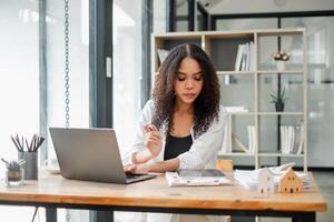 réel biens agent examine propriété les documents tandis que travail sur une portable dans une moderne bureau, avec miniature maison des modèles sur le tableau. photo